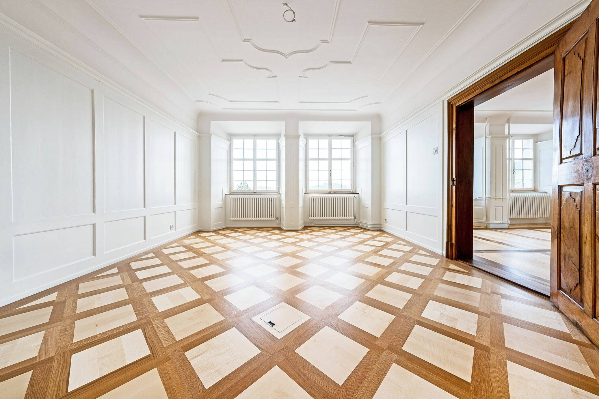 Zoom: Sicht in einen lichtdurchfluteten Raum mit Holzparkett und Stuckaturen an der Decke.