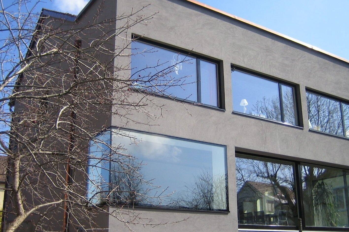 graue verputzte fassade eines mehrfamilienhauses mit grossen fenstern mit spiegelung von himmel, wolken und bäumen